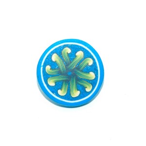 May include: A round, blue button with a white border and a central design of green and yellow curved lines. The button's design is symmetrical, with the lines radiating from the center.