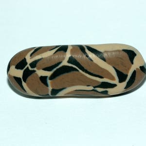 Puede incluir: Una pequeña cuenta oblonga con un patrón de camuflaje. La cuenta presenta una mezcla de formas orgánicas marrones, beige y negras. La cuenta es lisa y parece estar hecha de un material sólido.