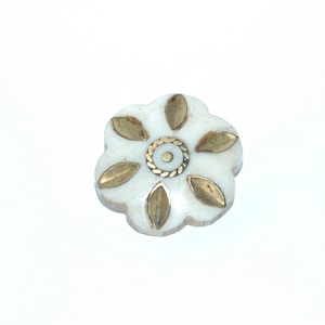 May include: A white, flower-shaped decorative button with six gold-colored leaf accents and a gold-colored center ring. The button is set against a white background.