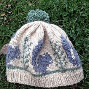 以下が含まれることがあります： クリーム色のニット帽に緑色のポンポンが付いている。帽子には青いリスと緑の葉の模様がある。