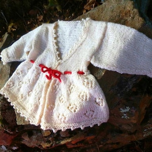 Può includere: Un cardigan bianco lavorato a maglia per neonati con un nastro rosso. Il cardigan ha un delicato motivo a pizzo e una scollatura a V.