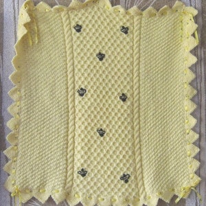 May include: A yellow knitted baby blanket with a honeycomb pattern and embroidered bees. The blanket has a scalloped edge and yellow ribbon ties.