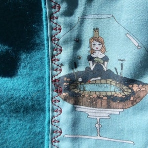 Pu&ograve; includere: Un tessuto turchese con un bordo decorativo e un disegno stampato. Il disegno presenta una principessa in una ciotola di vetro con uno stagno. Il bordo &egrave; cucito con filo rosso. Il tessuto &egrave; morbido e strutturato.