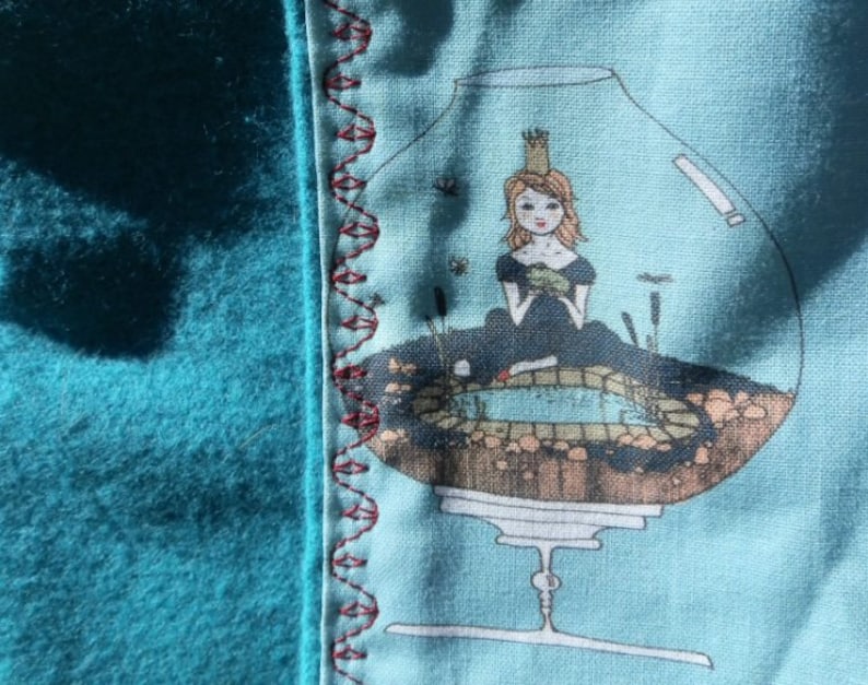 Pu&ograve; includere: Un tessuto turchese con un bordo decorativo e un disegno stampato. Il disegno presenta una principessa in una ciotola di vetro con uno stagno. Il bordo &egrave; cucito con filo rosso. Il tessuto &egrave; morbido e strutturato.