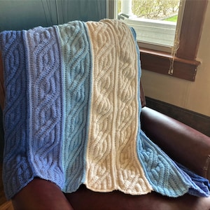 May include: A knitted throw blanket with a cable knit pattern in shades of blue and white. The blanket is draped over a brown leather armchair.