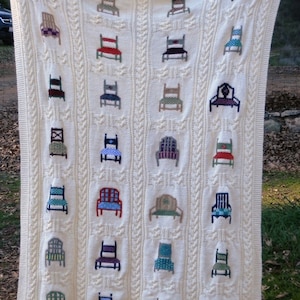 Puede incluir: Una manta de punto blanco con un patrón de punto de cable presenta un diseño repetitivo de sillas coloridas bordadas en la tela.