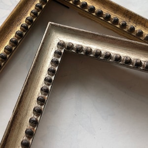 May include: Two ornate picture frames with decorative beading. One frame is gold with dark brown accents, the other is off-white with dark brown accents. The frames are angled, showing the corner details.