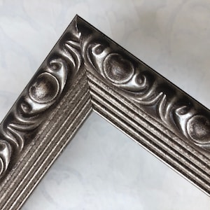 May include: Close-up of a silver picture frame with an ornate, raised design. The frame has a textured, metallic finish.