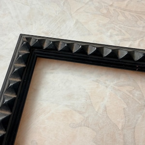 May include: Black picture frame with a decorative, textured edge. The frame's corner is visible, showcasing a series of pyramid-shaped details along the border. The frame is set against a neutral, patterned background.