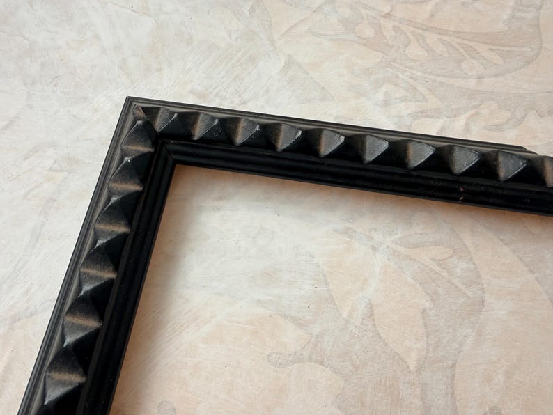 May include: Black picture frame with a decorative, textured edge. The frame's corner is visible, showcasing a series of pyramid-shaped details along the border. The frame is set against a neutral, patterned background.