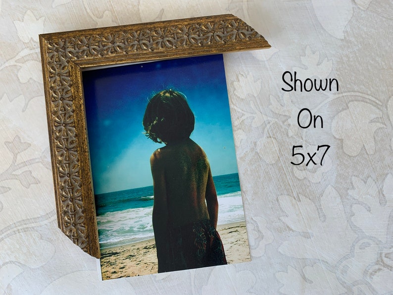 May include: A gold-toned picture frame with an ornate design. The frame is holding a photograph of a child standing on a beach, looking out at the ocean. The text "Shown On 5x7" is written on the white background behind the frame.