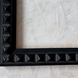 May include: A close-up of a black picture frame with a textured, geometric design along the edges. The frame has a matte finish and is set against a light-colored, patterned background.