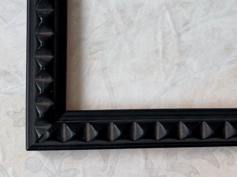 May include: A close-up of a black picture frame with a textured, geometric design along the edges. The frame has a matte finish and is set against a light-colored, patterned background.