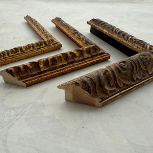 May include: Four ornate, antique picture frame corners in various shades of gold and brown. The frames have detailed, carved designs and are arranged on a light-colored surface, showcasing their intricate craftsmanship and vintage aesthetic.