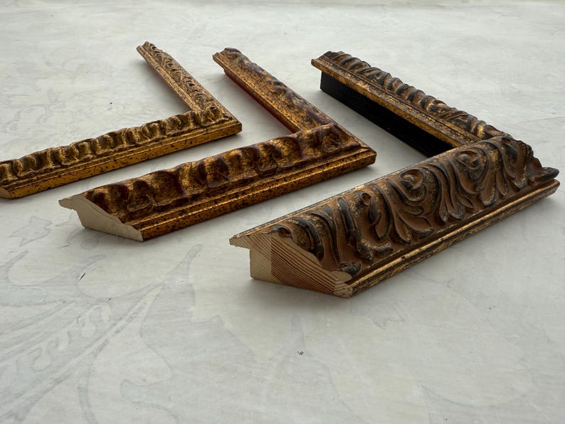 May include: Four ornate, antique picture frame corners in various shades of gold and brown. The frames have detailed, carved designs and are arranged on a light-colored surface, showcasing their intricate craftsmanship and vintage aesthetic.