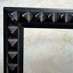 May include: Close-up of a black picture frame corner. The frame has a textured surface with repeating pyramid-shaped details along the edges. The background is a light-colored surface with a subtle floral pattern.
