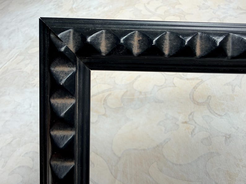 May include: Close-up of a black picture frame corner. The frame has a textured surface with repeating pyramid-shaped details along the edges. The background is a light-colored surface with a subtle floral pattern.