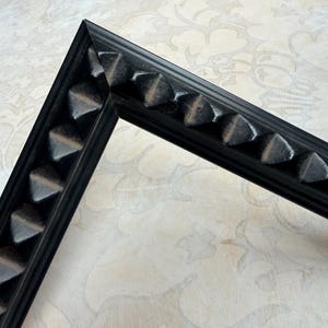 May include: A black picture frame corner with a repeating triangular design. The frame is made of a dark material and has a matte finish. The corner is at a 90-degree angle, showcasing the frame's construction and detail.