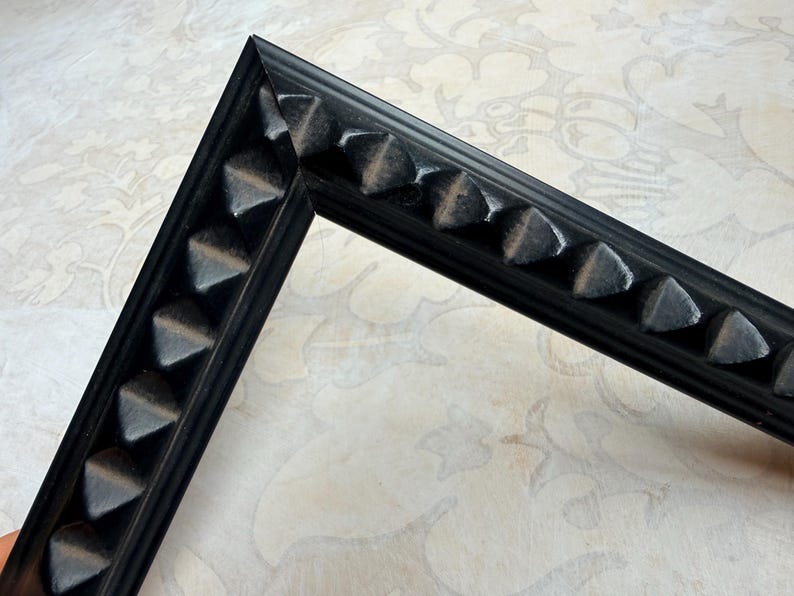 May include: A black picture frame corner with a repeating triangular design. The frame is made of a dark material and has a matte finish. The corner is at a 90-degree angle, showcasing the frame's construction and detail.