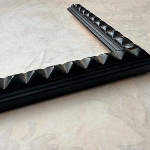 May include: A corner of a black picture frame with a textured, geometric design. The frame is angled, showcasing the detailed pyramid-shaped accents along its edges. The frame rests on a light-colored, patterned surface.