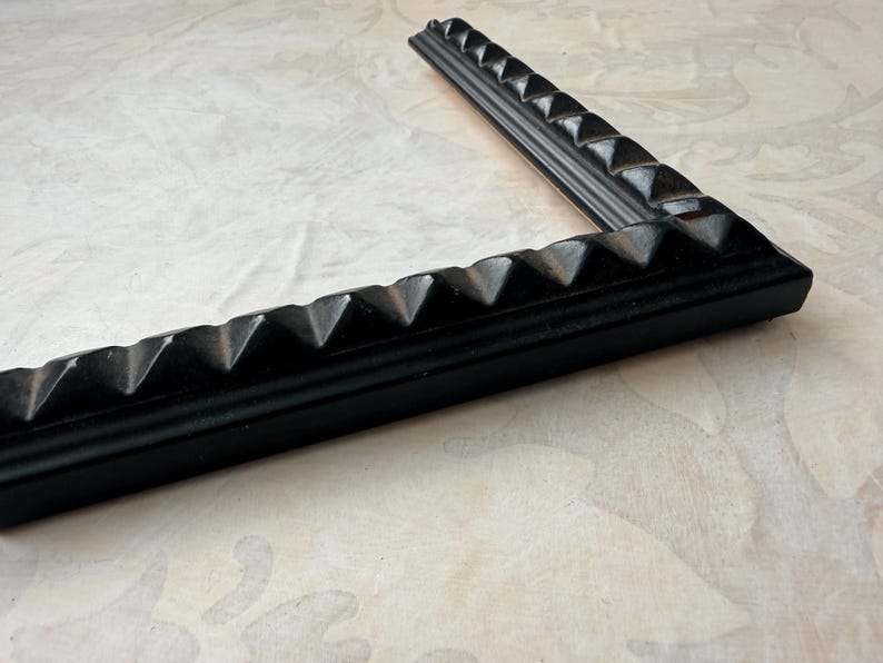 May include: A corner of a black picture frame with a textured, geometric design. The frame is angled, showcasing the detailed pyramid-shaped accents along its edges. The frame rests on a light-colored, patterned surface.