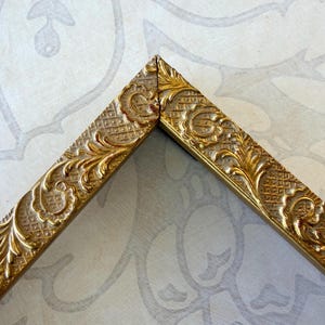 May include: A gold-colored picture frame corner section featuring an ornate, raised floral and leaf pattern set against a textured, cross-hatched background. The frame has a metallic finish with visible highlights on the embossed decorative elements.