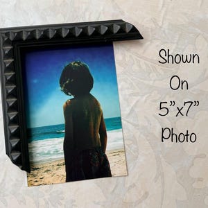 May include: Black picture frame with a textured, studded design. The frame holds a 5"x7" photo of a person on a beach. The photo shows a person looking out at the ocean under a blue sky. Text on the image reads "Shown On 5"x7" Photo."