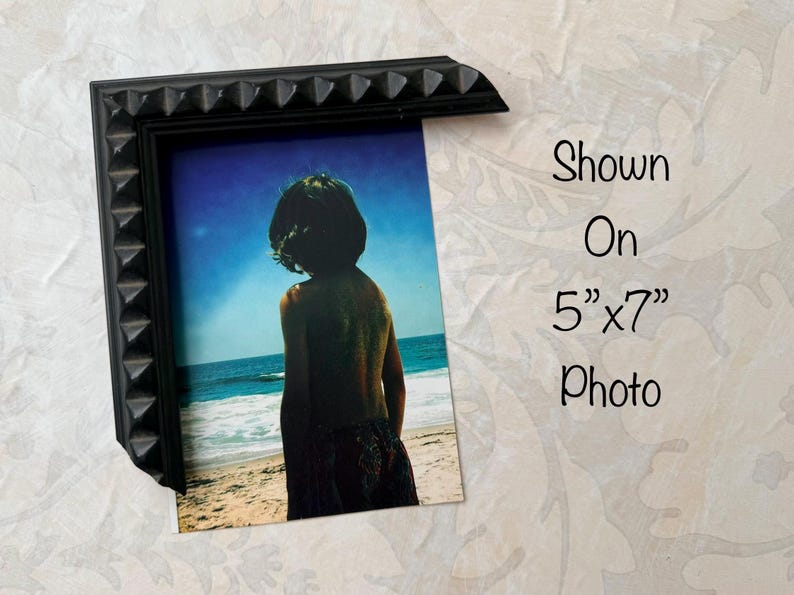 May include: Black picture frame with a textured, studded design. The frame holds a 5"x7" photo of a person on a beach. The photo shows a person looking out at the ocean under a blue sky. Text on the image reads "Shown On 5"x7" Photo."