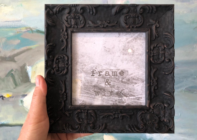 May include: A black ornate frame with a white background and the text "frame & wick" printed on it.