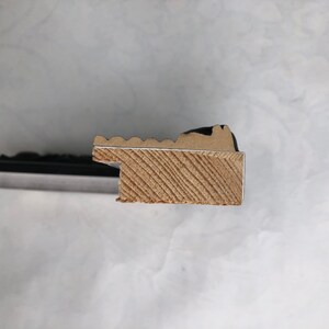 K&ouml;nnte beinhalten: Nahaufnahme der Seite eines schwarzen Bilderrahmens mit einem Holzkern. Der Holzkern hat eine hellbraune Farbe und ein diagonales Maserungsmuster.