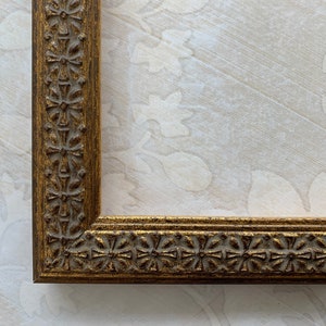 May include: A gold picture frame with a decorative, ornate design. The frame has a raised edge and a detailed pattern.