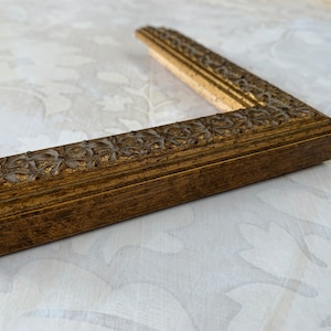 May include: A gold picture frame with a decorative, ornate design. The frame is made of wood and has a dark brown finish.