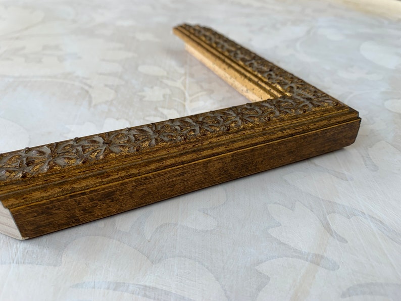 May include: A gold picture frame with a decorative, ornate design. The frame is made of wood and has a dark brown finish.