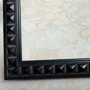 May include: A black picture frame with a geometric, pyramid-shaped design along the edges. The frame surrounds a light-colored background with a subtle floral pattern. The frame is made of a dark, possibly wooden material.
