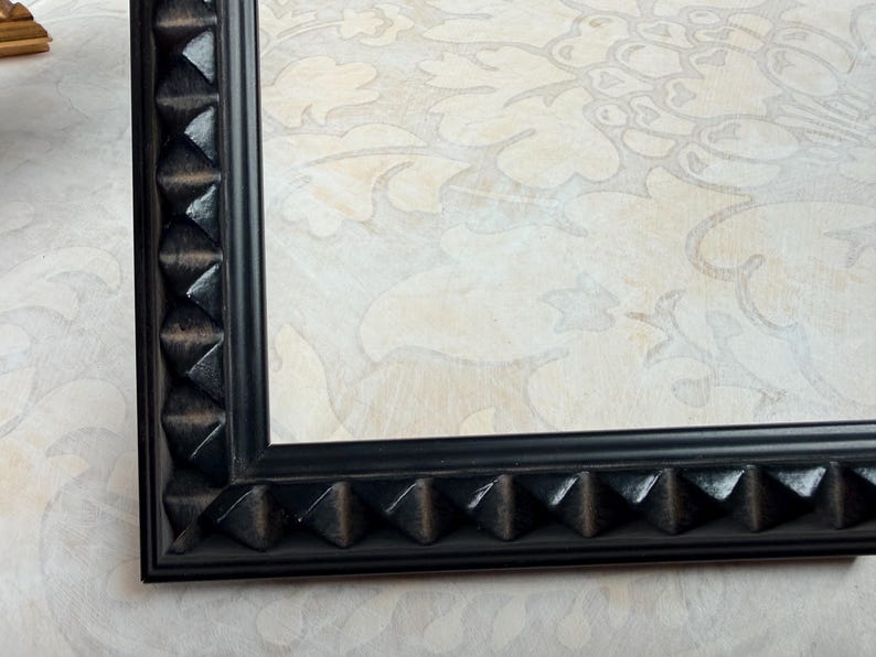 May include: A black picture frame with a geometric, pyramid-shaped design along the edges. The frame surrounds a light-colored background with a subtle floral pattern. The frame is made of a dark, possibly wooden material.