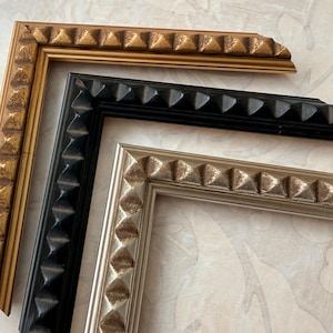 May include: Close-up of three picture frame corners in gold, black, and silver. Each frame has a repeating pyramid design along the edges. The frames are arranged on a patterned, light-colored surface.
