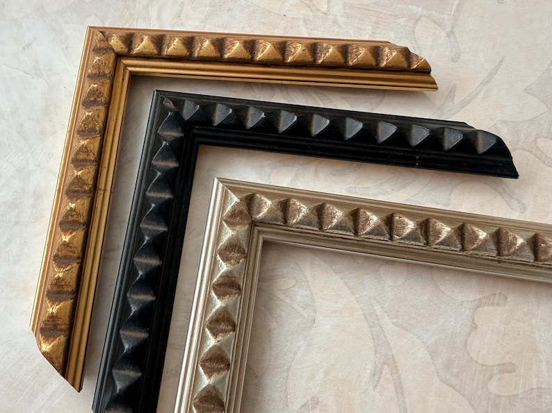 May include: Close-up of three picture frame corners in gold, black, and silver. Each frame has a repeating pyramid design along the edges. The frames are arranged on a patterned, light-colored surface.