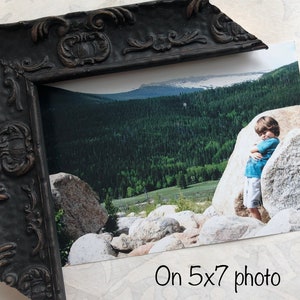 May include: A black ornate picture frame with a 5x7 inch photo of a child standing in a mountainous landscape. The photo shows a child standing next to a large rock with a green forest and a blue sky in the background.