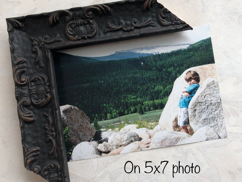 May include: A black ornate picture frame with a 5x7 inch photo of a child standing in a mountainous landscape. The photo shows a child standing next to a large rock with a green forest and a blue sky in the background.