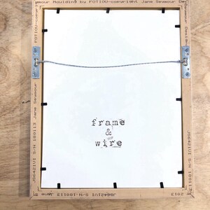 May include: Back of a framed artwork with a light brown wooden frame. The white backing has the words "frame & wire" printed on it. Metal brackets and wire are attached for hanging. The frame has text that reads "Jane Seymour Designs".