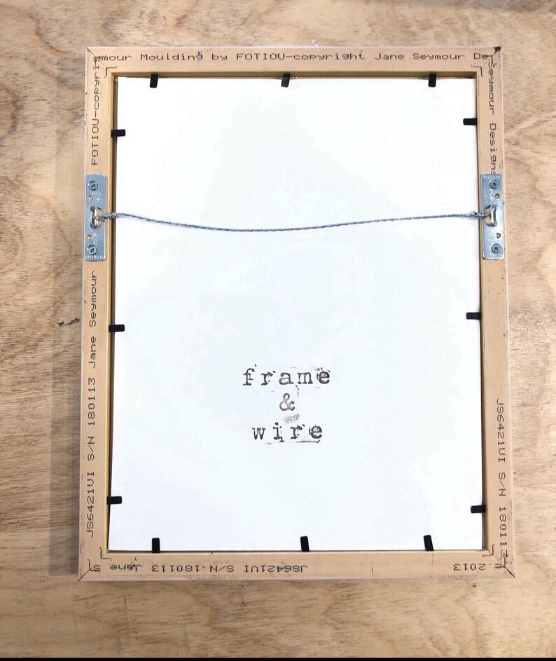 May include: Back of a framed artwork with a light brown wooden frame. The white backing has the words "frame & wire" printed on it. Metal brackets and wire are attached for hanging. The frame has text that reads "Jane Seymour Designs".