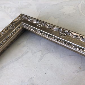 May include: Close-up of a silver-colored picture frame corner. The frame has ornate, scrolling details along the edges. The frame is made of a metallic material and has a vintage or antique aesthetic. The frame is likely for displaying photos or artwork.