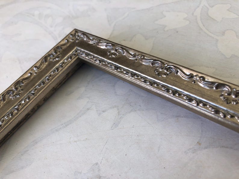 May include: Close-up of a silver-colored picture frame corner. The frame has ornate, scrolling details along the edges. The frame is made of a metallic material and has a vintage or antique aesthetic. The frame is likely for displaying photos or artwork.