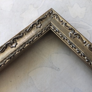 May include: A corner of a gold-colored picture frame with ornate, raised floral and scrollwork detailing. The frame has a slightly distressed finish, adding to its vintage aesthetic. The frame's design is classic and elegant.