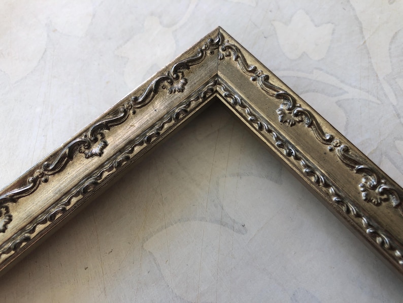 May include: A corner of a gold-colored picture frame with ornate, raised floral and scrollwork detailing. The frame has a slightly distressed finish, adding to its vintage aesthetic. The frame's design is classic and elegant.