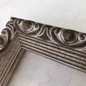 May include: A silver picture frame with a decorative, ornate border. The frame has a distressed, aged look.