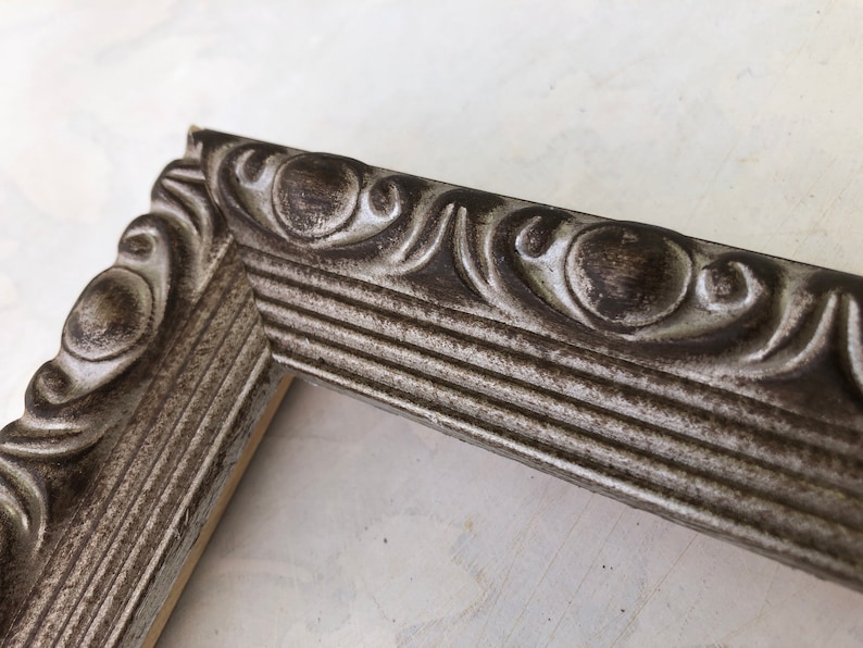 May include: A silver picture frame with a decorative, ornate border. The frame has a distressed, aged look.