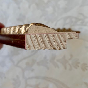 May include: Close-up of the side of a gold picture frame. The frame is made of wood and has a decorative, ornate design. The frame is painted gold and has a red-brown inner edge.