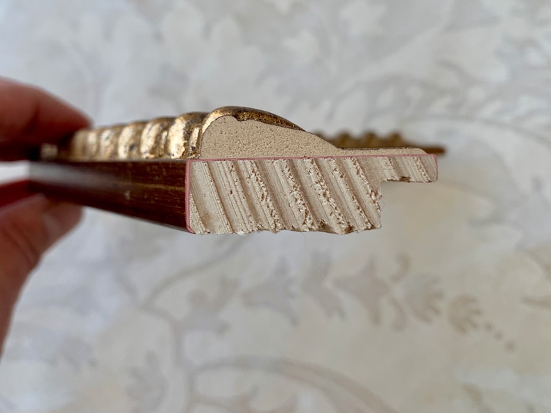 May include: Close-up of the side of a gold picture frame. The frame is made of wood and has a decorative, ornate design. The frame is painted gold and has a red-brown inner edge.
