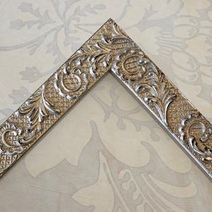 May include: A silver-toned picture frame corner featuring an intricate embossed pattern of scrolling acanthus leaves and a textured cross-hatched grid design. The metallic finish has a reflective surface with dark highlights in the recessed areas.